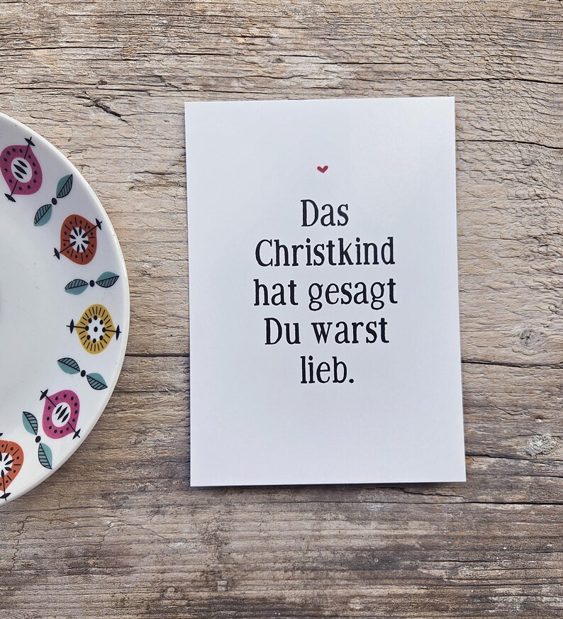 May include: A white rectangular greeting card with black text that reads Das Christkind hat gesagt Du warst lieb and a small red heart icon above the text, positioned next to a white plate with a colorful geometric pattern.