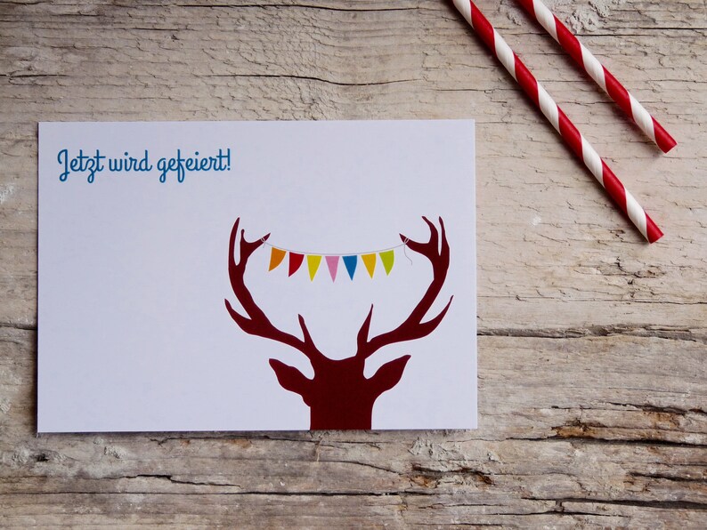 May include: A white card with the text "Jetzt wird gefeiert!" in blue. A red deer silhouette with antlers is on the card. A colorful string of bunting hangs from the antlers.