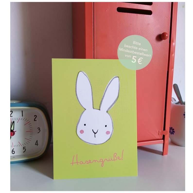 Peut inclure: Une carte verte avec une illustration de lapin blanc et le texte "Hasengrube!" en rose. Un petit cercle vert avec le texte "Bitte beachte einen Mindestbestellwert von 5&euro;" est dans le coin sup&eacute;rieur droit.