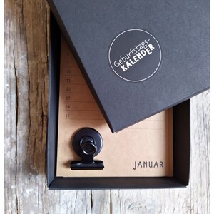 May include: A black box with a white circle sticker that says "Geburtstagskalender" on the top. The box is open and a brown paper calendar with a black clip is inside. The calendar says "Januar" at the bottom.