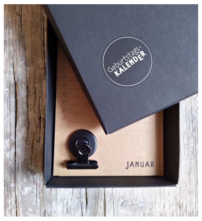 May include: A black box with a white circle sticker that says "Geburtstagskalender" on the top. The box is open and a brown paper calendar with a black clip is inside. The calendar says "Januar" at the bottom.