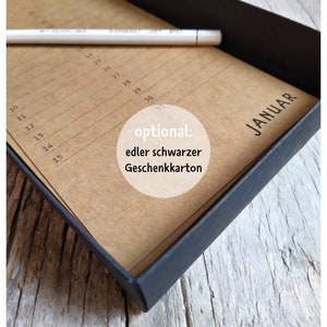 May include: A brown paper calendar with the month of January printed on it. The calendar is in a black box. The text "optional: edler schwarzer Geschenkkarton" is printed on the calendar.