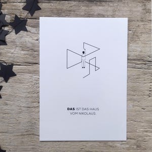 May include: A white rectangular card featuring a black line drawing of a person drawing a house shape with the text DAS IST DAS HAUS VOM NIKOLAUS printed in black below the illustration. The card is positioned next to black paper stars.