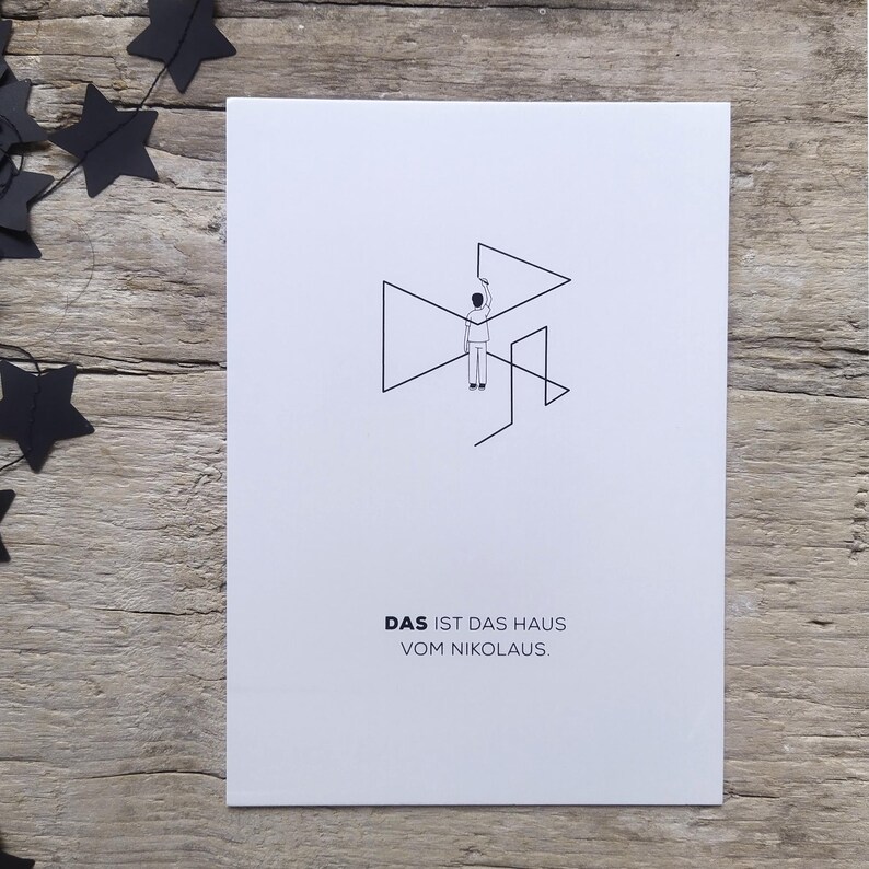 May include: A white rectangular card featuring a black line drawing of a person drawing a house shape with the text DAS IST DAS HAUS VOM NIKOLAUS printed in black below the illustration. The card is positioned next to black paper stars.