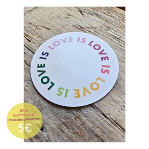 May include: A round white sticker with the words "LOVE IS LOVE" in a rainbow of colors. The sticker is on a wooden surface. A yellow circle with German text and the number 5 is in the lower left corner.