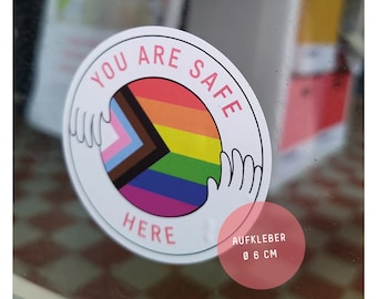 Könnte beinhalten: Ein runder weißer Aufkleber mit dem Text "You Are Safe Here" in rosa Schrift. Der Aufkleber zeigt eine Regenbogen-Pride-Flagge mit einem schwarzen, braunen und hellblauen Dreieck in der Mitte. Zwei rosa Hände befinden sich auf beiden Seiten der Flagge und bilden eine schützende Geste. Der Aufkleber enthält auch den Text "AUFKLEBER Ø 6 CM" in rosa Schrift.