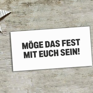 May include: A rectangular white card featuring the black printed text M&Ouml;GE DAS FEST MIT EUCH SEIN! in a bold sans-serif font. The card is positioned on a light-colored wooden surface next to a folded paper star made from printed newspaper.