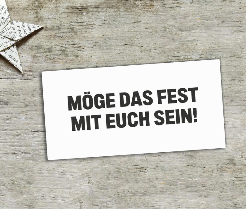 May include: A rectangular white card featuring the black printed text M&Ouml;GE DAS FEST MIT EUCH SEIN! in a bold sans-serif font. The card is positioned on a light-colored wooden surface next to a folded paper star made from printed newspaper.
