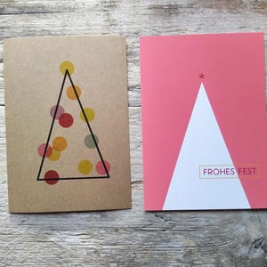 May include: Two rectangular greeting cards. The left card is brown with a black triangle outline containing translucent yellow, orange, and red circles. The right card is pink with a white triangle and the text FROHES FEST in a gold box.