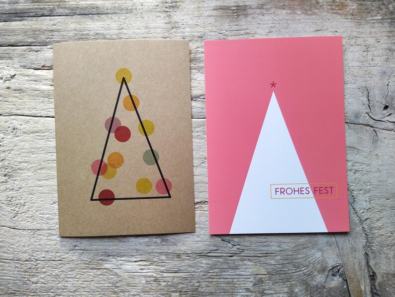 May include: Two rectangular greeting cards. The left card is brown with a black triangle outline containing translucent yellow, orange, and red circles. The right card is pink with a white triangle and the text FROHES FEST in a gold box.