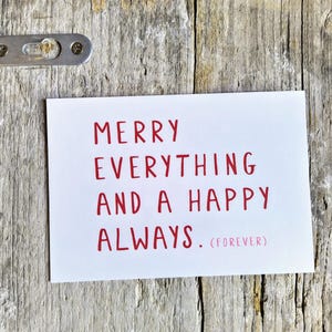 May include: A rectangular white greeting card featuring the red text MERRY EVERYTHING AND A HAPPY ALWAYS. (FOREVER) printed in a simple sans-serif font. The card is positioned against a weathered, light-colored wooden surface with a metal bracket.