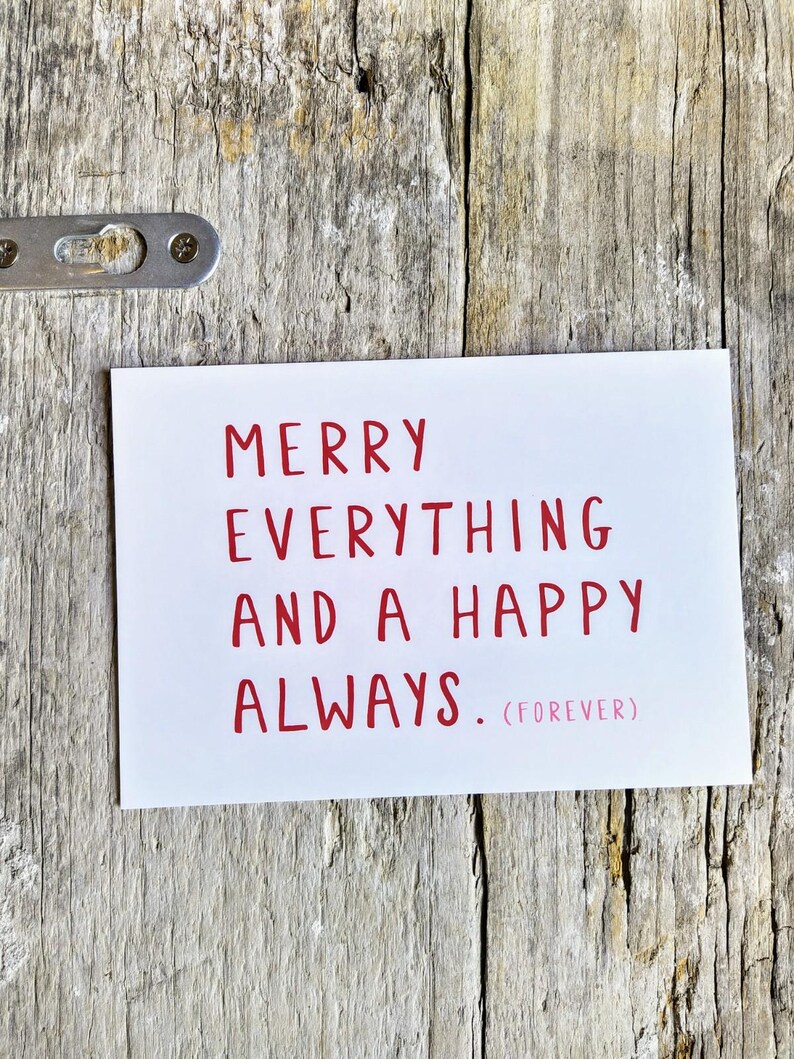 May include: A rectangular white greeting card featuring the red text MERRY EVERYTHING AND A HAPPY ALWAYS. (FOREVER) printed in a simple sans-serif font. The card is positioned against a weathered, light-colored wooden surface with a metal bracket.