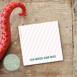 Könnte beinhalten: Eine quadratische Karte mit diagonalen rosa und weißen Streifen, mit dem Text "ICH MUSS GAR NIX!" in Grün. Ein roter Tentakel und ein grüner Kreis mit Text sind ebenfalls auf einer Holzoberfläche zu sehen.