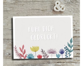 May include: A light gray card with the text "Fühl dich gedrückt!" in white lettering. The card is decorated with a border of colorful flowers and leaves.