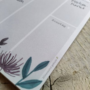 May include: A white weekly planner with a floral design at the bottom. The planner has a grid layout with days of the week labelled in German. The text "klimaneutral gedruckt" is printed on the bottom right corner.