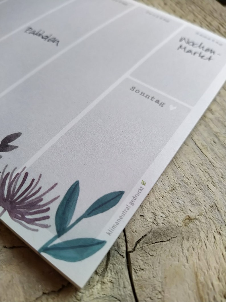 May include: A white weekly planner with a floral design at the bottom. The planner has a grid layout with days of the week labelled in German. The text "klimaneutral gedruckt" is printed on the bottom right corner.