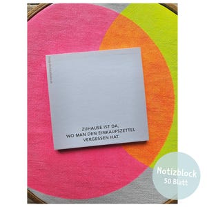 May include: A square notepad with white paper featuring the black printed German text: ZUHAUSE IST DA, WO MAN DEN EINKAUFSZETTEL VERGESSEN HAT. The notepad is placed on a circular wooden embroidery hoop frame with a vibrant pink, orange, and lime green background.
