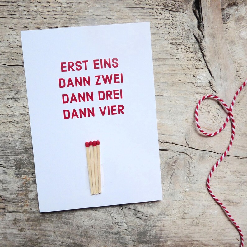 May include: A white rectangular greeting card featuring the red printed text ERST EINS DANN ZWEI DANN DREI DANN VIER. Below the text, four wooden matchsticks with red tips are attached to the card in a vertical row, aligned side by side.