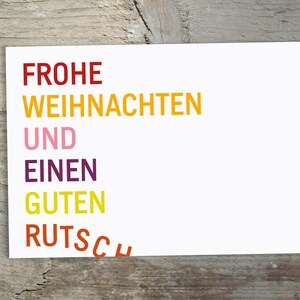 May include: A rectangular white greeting card featuring the German text FROHE WEIHNACHTEN UND EINEN GUTEN RUTSCH printed in stacked lines. The words are colored in red, yellow, pink, purple, yellow, and orange, with the final word slightly curved.