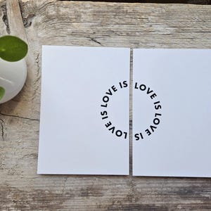 May include: Two white cards with the words "LOVE IS" arranged in a circle. The cards are placed on a weathered wooden surface. A small white pot with a green plant is visible in the upper left corner.