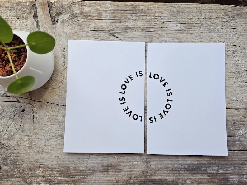 May include: Two white cards with the words "LOVE IS" arranged in a circle. The cards are placed on a weathered wooden surface. A small white pot with a green plant is visible in the upper left corner.