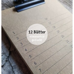 May include: A brown clipboard with a black clip at the top. The clipboard has 12 lined pages and the text "12 Bl&auml;tter klimaneutral gedruckt" is printed on the front.