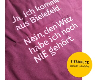 May include: A maroon tote bag with white text that reads "Ja, ich komme aus Bielefeld. Nein, den Witz habe ich noch NIE gehört." The bottom of the bag has a yellow label that reads "SIEBDRUCK gedruckt in Bielefeld".