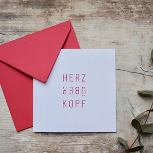 May include: A white card with the German phrase "Herz &uuml;ber Kopf" printed in red. The card is sitting on a wooden surface with a red envelope and a sprig of eucalyptus.