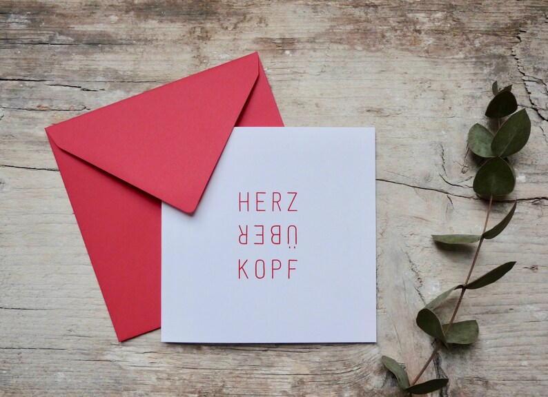 May include: A white card with the German phrase "Herz &uuml;ber Kopf" printed in red. The card is sitting on a wooden surface with a red envelope and a sprig of eucalyptus.