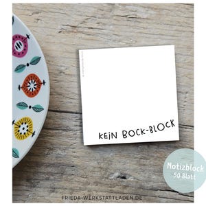 May include: A white notepad with the words "KEIN BOCK-BLOCK" in black, and a decorative plate with colourful floral designs. The notepad is on a wooden surface.