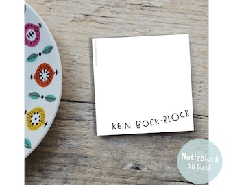 May include: A white notepad with the words "KEIN BOCK-BLOCK" in black, and a blue circle with the words "Notizblock 50 Blatt". The notepad is on a wooden surface, with a decorative plate in the background.