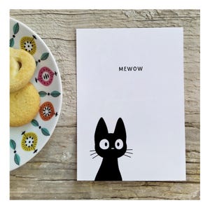 May include: A white rectangular greeting card featuring a black cat illustration at the bottom and the text MEWOW printed in black near the top, positioned next to a white plate with a colorful floral pattern containing two round cookies.