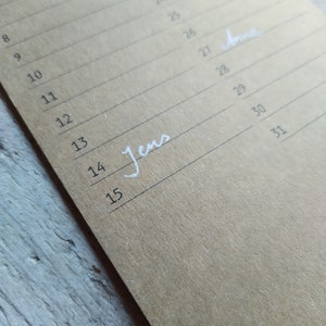 May include: A close-up of a brown paper calendar with the numbers 8 through 15 and 21 through 31 written in black ink. The word "Jens" is written in white ink on the line for the 14th and the word "Anne" is written in white ink on the line for the 27th.