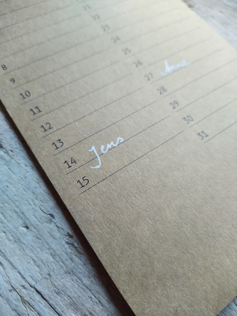 May include: A close-up of a brown paper calendar with the numbers 8 through 15 and 21 through 31 written in black ink. The word "Jens" is written in white ink on the line for the 14th and the word "Anne" is written in white ink on the line for the 27th.