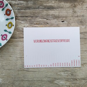 May include: A white rectangular card featuring the red text VIERUNDZWANZIGTAGEVORFREUDE and a numbered countdown list from eins to vierundzwanzig at the bottom, positioned next to a white plate with a colorful geometric pattern of pink, orange, and yellow.