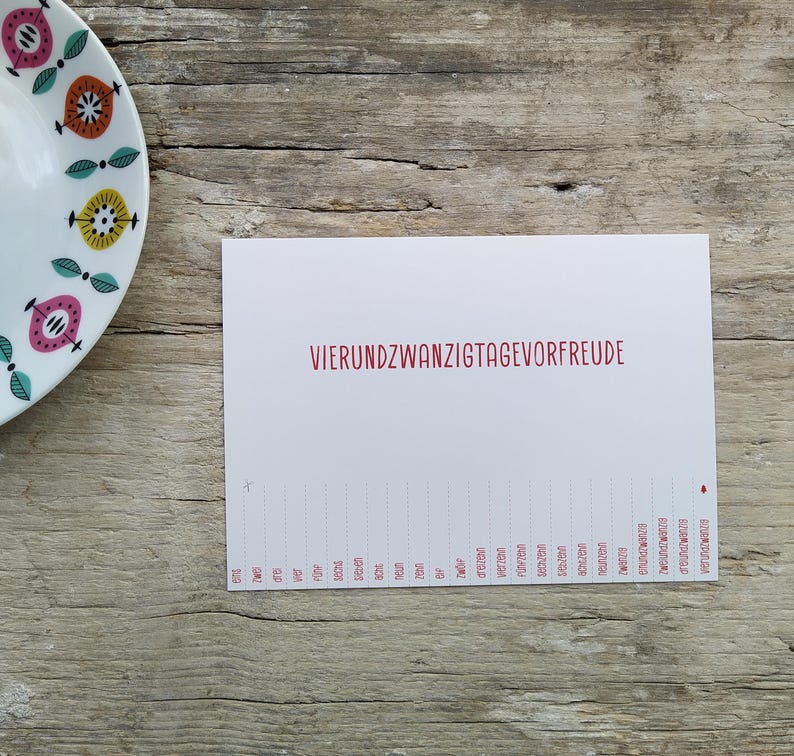 May include: A white rectangular card featuring the red text VIERUNDZWANZIGTAGEVORFREUDE and a numbered countdown list from eins to vierundzwanzig at the bottom, positioned next to a white plate with a colorful geometric pattern of pink, orange, and yellow.