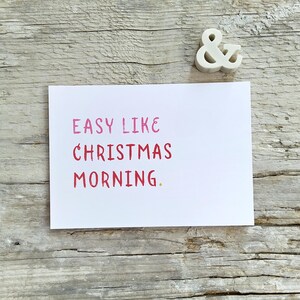 May include: A white rectangular greeting card featuring the pink and red text "EASY LIKE CHRISTMAS MORNING." positioned above a small white ampersand symbol. The card has a clean, simple design with bold, sans-serif lettering on a plain background.