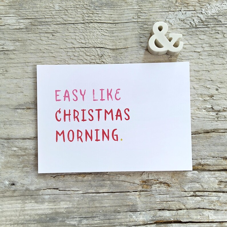 May include: A white rectangular greeting card featuring the pink and red text "EASY LIKE CHRISTMAS MORNING." positioned above a small white ampersand symbol. The card has a clean, simple design with bold, sans-serif lettering on a plain background.