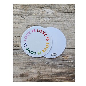 May include: Two white circular coasters with the phrase "LOVE IS LOVE" in rainbow colors. One coaster is centered, and the other is partially visible. The coasters are on a wooden surface.