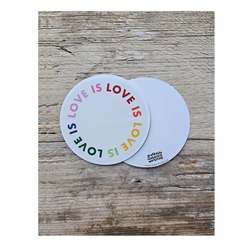 May include: Two white circular coasters with the phrase "LOVE IS LOVE" in rainbow colors. One coaster is centered, and the other is partially visible. The coasters are on a wooden surface.