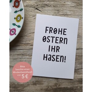 Peut inclure: Carte blanche avec du texte noir qui dit "Frohe Ostern Ihr Hasen!" La carte a un autocollant rose et blanc en bas qui dit "Bitte beachte einen Mindestbestellwert von 5&euro;".