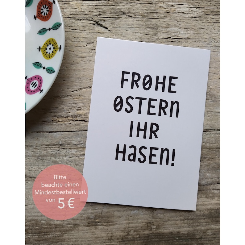 Peut inclure: Carte blanche avec du texte noir qui dit "Frohe Ostern Ihr Hasen!" La carte a un autocollant rose et blanc en bas qui dit "Bitte beachte einen Mindestbestellwert von 5&euro;".