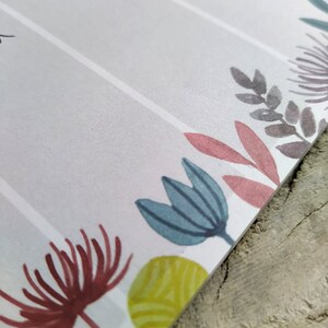 May include: A white planner with a floral design. The planner has a grey, pink, blue, and green floral pattern. The planner has a white background and the floral design is on the bottom edge of the planner.