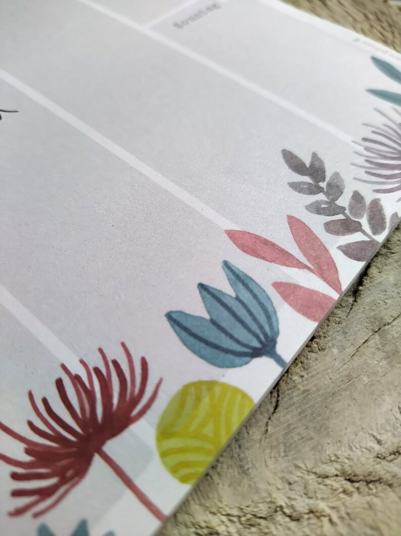 May include: A white planner with a floral design. The planner has a grey, pink, blue, and green floral pattern. The planner has a white background and the floral design is on the bottom edge of the planner.