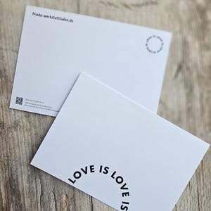May include: Two white postcards with the text "LOVE IS LOVE" in a semi-circular arrangement. One postcard is angled towards the viewer, while the other is in the background. The postcards are on a wooden surface.