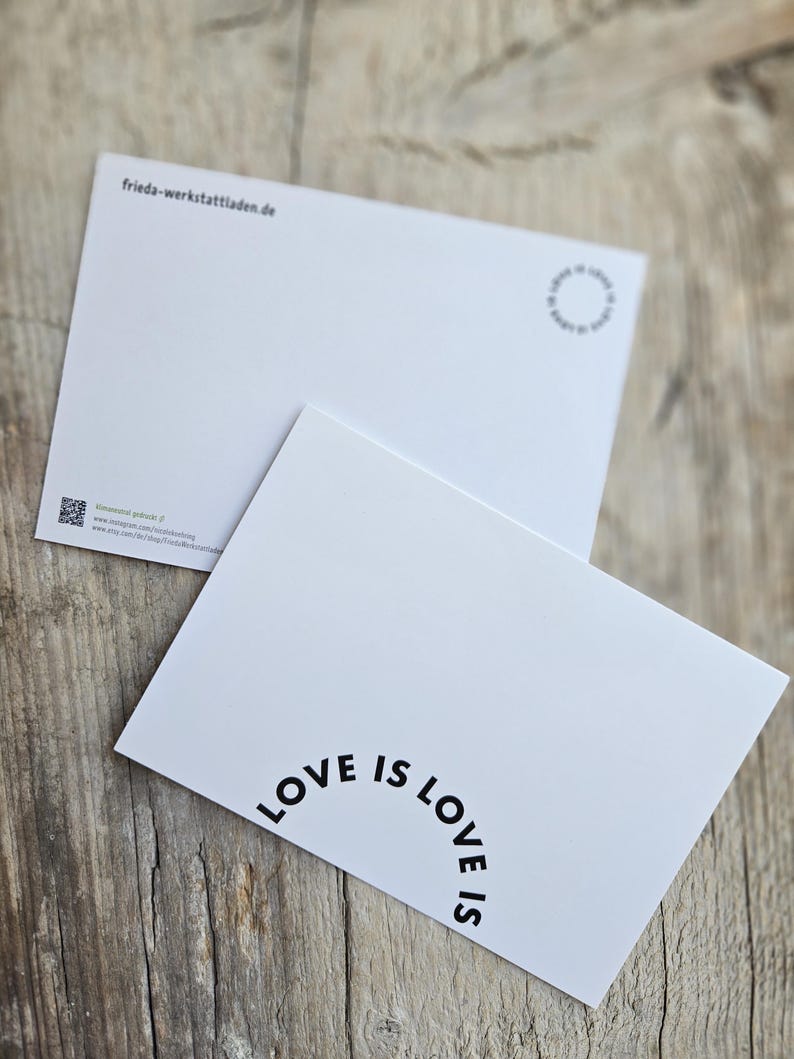 May include: Two white postcards with the text "LOVE IS LOVE" in a semi-circular arrangement. One postcard is angled towards the viewer, while the other is in the background. The postcards are on a wooden surface.