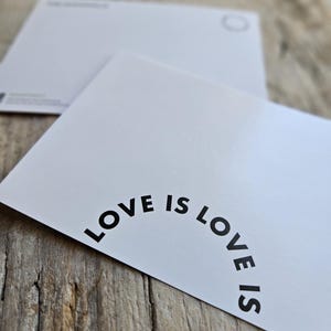 May include: Two white postcards with rounded corners are displayed on a wooden surface. The front postcard has the phrase "LOVE IS LOVE IS" printed in black, curved along the top edge. The second postcard is visible in the background.