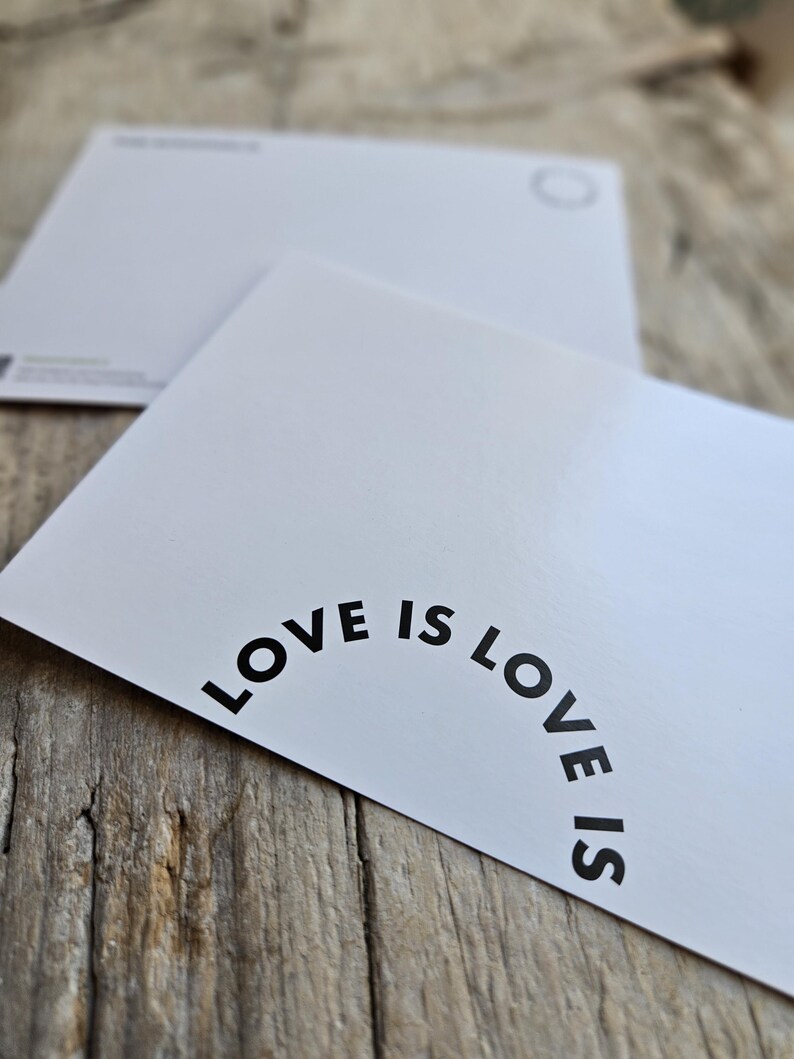 May include: Two white postcards with rounded corners are displayed on a wooden surface. The front postcard has the phrase "LOVE IS LOVE IS" printed in black, curved along the top edge. The second postcard is visible in the background.
