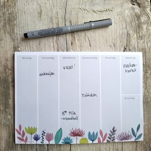 May include: A weekly planner with a grey background and a floral border. The days of the week are listed at the top: Monday, Tuesday, Wednesday, Thursday, Friday, Saturday, and Sunday. The planner has handwritten notes for each day, including "einkaufen", "FREI!", "Pabhden", "Wochen-Markt", and "18: Mila - Handball".