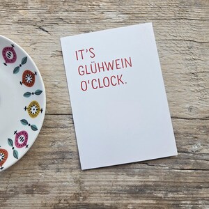 May include: A white rectangular greeting card with the red text IT'S GL&Uuml;HWEIN O'CLOCK. positioned next to a white ceramic plate featuring a repeating pattern of stylized pink, orange, and yellow circular shapes with small green leaves.
